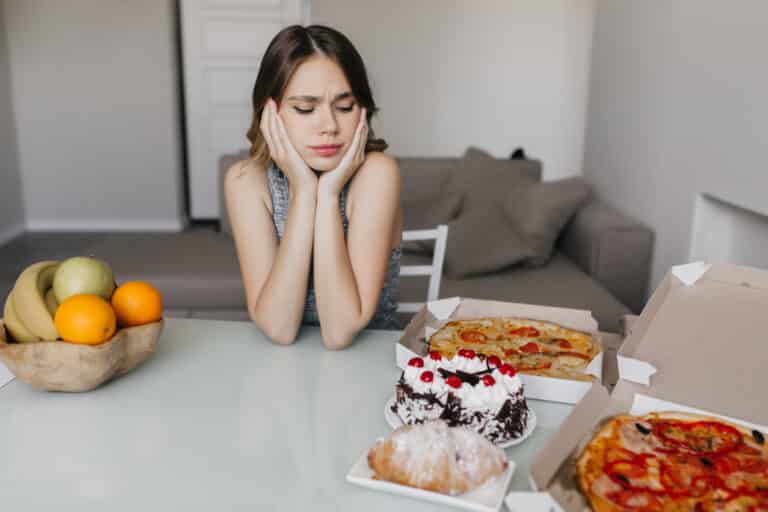 Sad curly woman looking at cake during diet. Blonde gorgeous female model posing with fruits and pizza